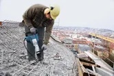 Asmuo, dėvintis saugos įrangą, griovimo plaktuku skaldo betoną ant stogo.