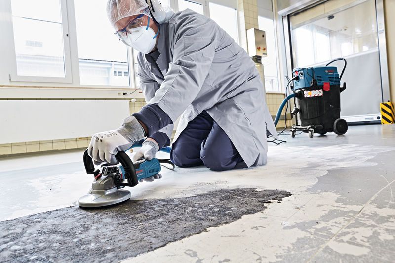 Asmuo, dėvintis apsaugos priemones, elektriniu įrankiu šlifuoja betonines grindis.