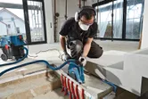 Asmuo, dėvintis apsaugos priemones, sienų sriegtuvu pjauna betoną šalia atvirų vamzdžių.