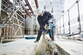 Asmuo, dėvintis saugos įrangą, statybvietėje naudoja sukamąjį plaktuką betonui skaldyti.