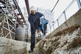 Asmuo, dėvintis saugos įrangą, statybvietėje naudoja sukamąjį plaktuką betonui ardyti.