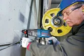 Asmuo, dėvintis apsaugos įrangą, gręžia betoną sukamuoju plaktuku šalia geltono skriemulio.