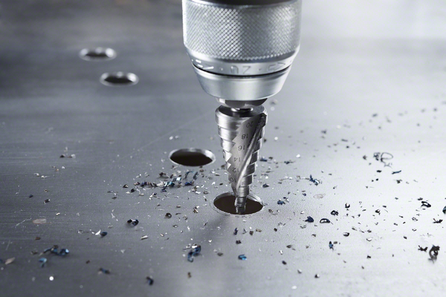 Laiptinis grąžtas, gręžiantis skylę metaliniame lakšte su išsklaidytomis drožlėmis.