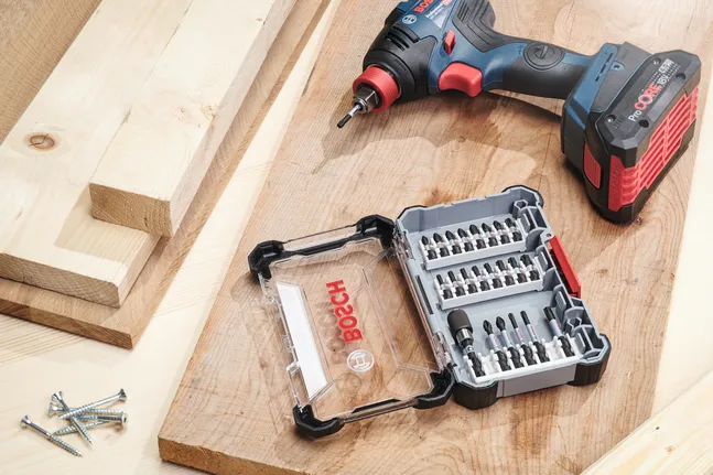 Akumuliatorinis gręžtuvas šalia grąžtų ir varžtų rinkinio ant medinio darbastalio.