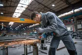 Asmuo, dėvintis apsaugos priemones, gamykloje šlifuoja metalą akumuliatoriniu kampiniu šlifuokliu.