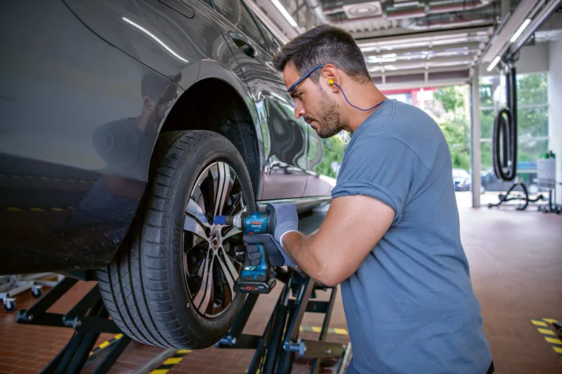 Asmuo, dėvintis apsaugos įrangą, smūginiu veržliarakčiu priveržia automobilio rato varžtus.
