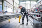 Apsauginę įrangą dėvintis asmuo dirbtuvėse šlifuoja metalinius kraštus ant perforuotos sijos.
