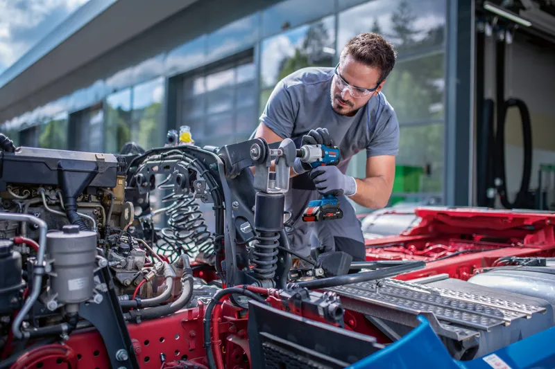 Asmuo, dėvintis apsaugos įrangą, smūginiu veržliasukiu priveržia automobilio važiuoklės varžtą.