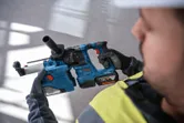 Asmuo, dėvintis apsaugos įrangą, laiko akumuliatorinį perforatorių-gręžtuvą.