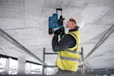 Asmuo, dėvintis apsaugos įrangą, gręžia betonines lubas elektriniu įrankiu.