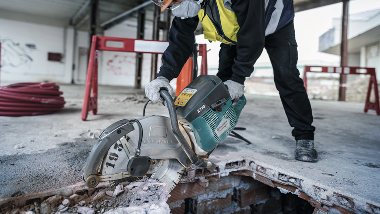 Asmuo, dėvintis saugos įrangą, statybvietėje pjauna betoną elektriniu pjūklu.