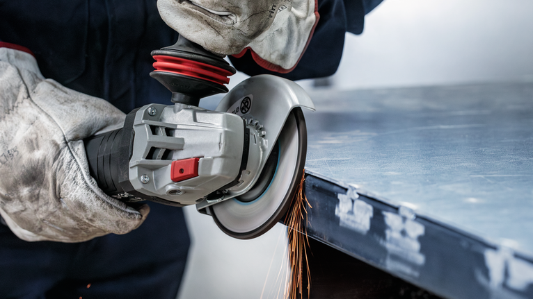 Apsauginę įrangą dėvintis asmuo elektriniu įrankiu šlifuoja metalinio lakšto kraštą.