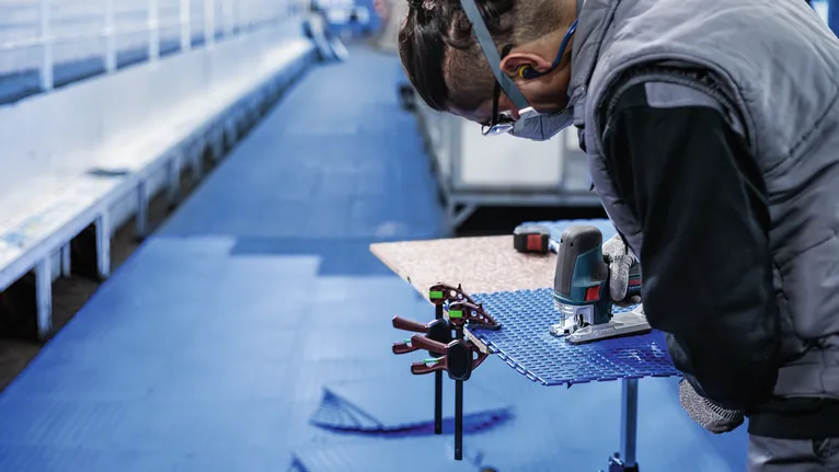 Asmuo, dėvintis saugos įrangą, elektriniu įrankiu pjausto mėlyną lakštą ant darbastalio.