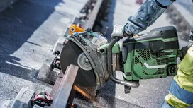 Asmuo, dėvintis apsaugos įrangą, rankiniu elektriniu pjūklu pjauna metalinį bėgelį.