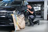 Asmuo, dėvintis apsaugos įrangą, autoservise valo automobilio sparną poliruokliu.