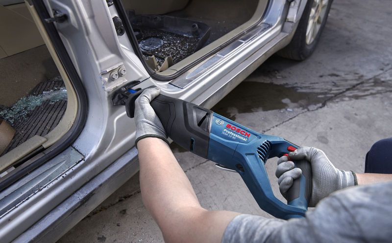 Asmuo, dėvintis apsaugos įrangą, slankiojančiu pjūklu pjausto metalą automobilio durelių rėme.