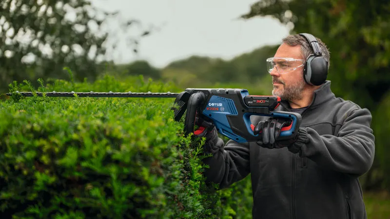 Asmuo, dėvintis apsaugos įrangą, kerpa gyvatvores elektrinėmis gyvatvorių žirklėmis.