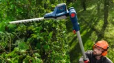 Asmuo, dėvintis apsaugos įrangą, karpo aukštas gyvatvores naudodamas akumuliatorines teleskopines gyvatvorių žirkles.