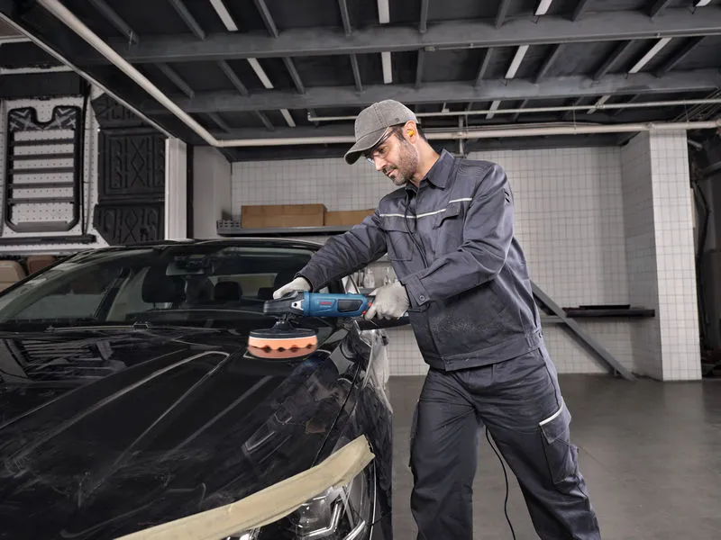Asmuo, dėvintis apsaugos įrangą, elektriniu įrankiu poliruoja automobilio kapotą.