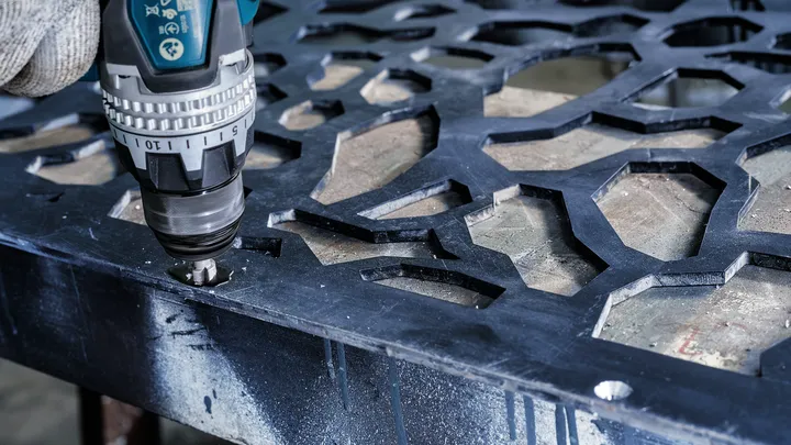 Asmuo, dėvintis apsaugos priemones, naudoja akumuliatorinį gręžtuvą, kad išgręžtų skylę metaliniame lakšte.