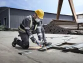 Asmuo, dėvintis apsaugos priemones, lauke šlifuoja metalines sijas kampiniu šlifuokliu.