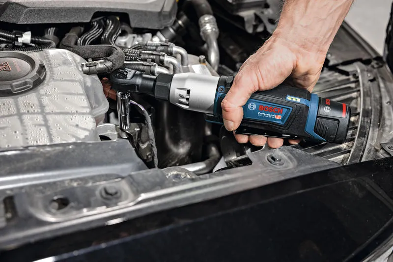 Žmogus naudoja akumuliatorinį reketinį veržliaraktį, kad priveržtų varžtą automobilio variklio viduje.