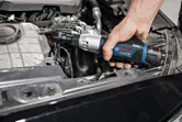 Žmogus naudoja akumuliatorinį reketinį veržliaraktį, kad priveržtų varžtą automobilio variklio viduje.