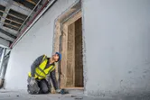 Asmuo, dėvintis apsaugos įrangą, lazeriniu matuokliu matuoja durų angą.