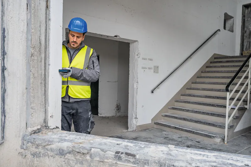 Asmuo, dėvintis apsaugos įrangą, lazeriniu matuokliu matuoja pastato angą.