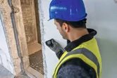 Asmuo, dėvintis apsaugos įrangą, matuoja sieną lazeriniu matuokliu.