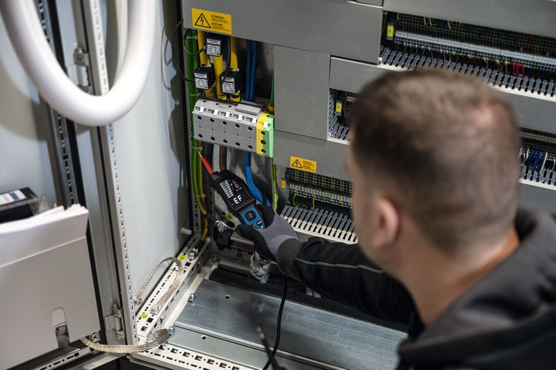Asmuo, dėvintis apsaugos įrangą, naudoja įtampos matuoklį elektros skydinės laidams matuoti.