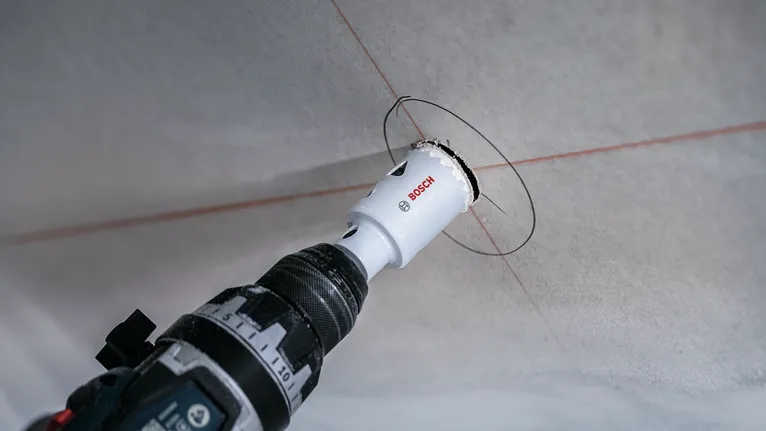 Elektrinis gręžtuvas su skylių pjūklo priedu, pjaustantis apvalią angą lubose.