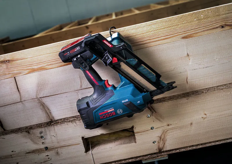 Akumuliatorinis karkasų vinių kalimo įrankis, stovintis ant medinio rėmo statybvietėje.