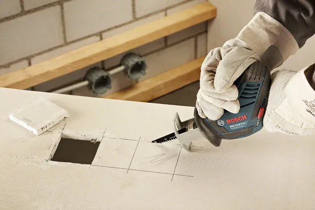 Apsauginęs įrangą darbuotojas slankiojančiu pjūklu išpjauna kvadratinę skylę gipso kartone.