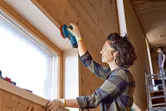 Persona, kas valkā drošības aprīkojumu, slīpē koka loga rāmi ar akumulatora ekscentra slīpmašīnu.