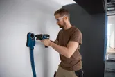 Persona, kas valkā drošības aprīkojumu, izmanto urbi un FlexiClick adapteri pie sienas.