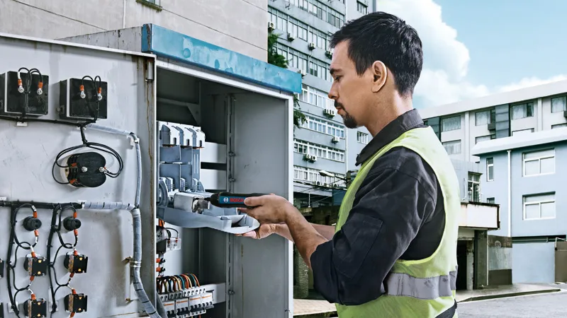 Persona, kas valkā drošības aprīkojumu, izmanto akumulatora skrūvgriezi uz elektrības paneļa.