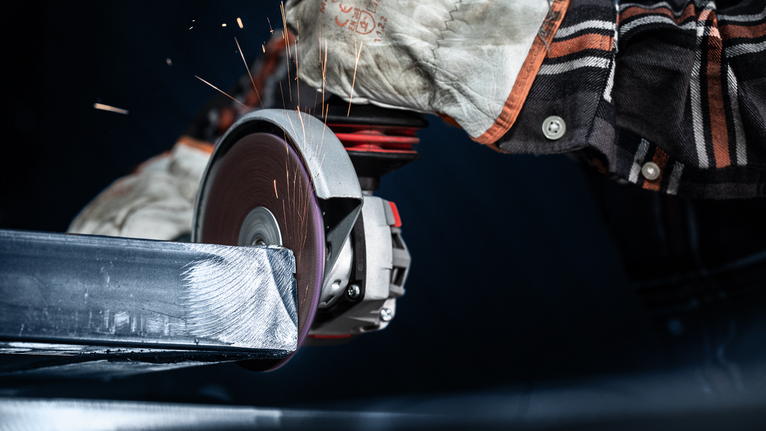 Persona, kas valkā drošības aprīkojumu, slīpē metāla siju ar elektroinstrumentu.