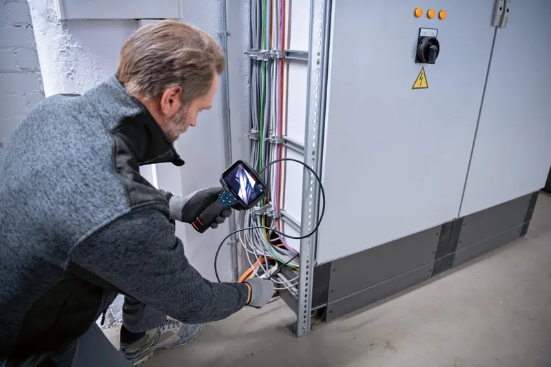 Persona, kas valkā drošības aprīkojumu, pārbauda elektroinstalāciju ar pārbaudes kameru.