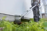 Persona, kas valkā drošības aprīkojumu, pļauj zāli gar sienu, izmantojot akumulatora zāles trimmeri.