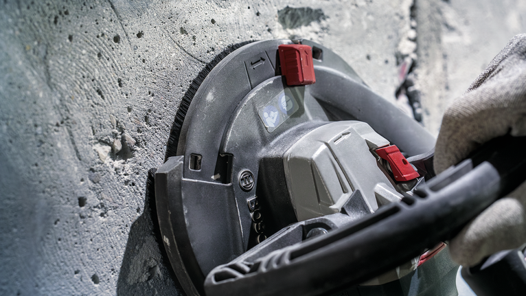 Persona drošības aprīkojumā slīpē betona sienu ar elektroinstrumentu.