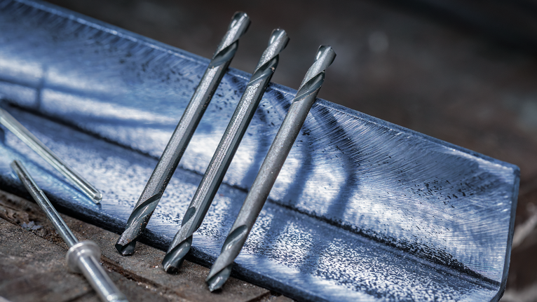 Trīs metāla urbju uzgaļi balstās uz zila tērauda leņķa uz darbagalda.