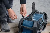 Persona, kas valkā drošības aprīkojumu, pievieno uzlādes kabeli izturīgam darba vietas radio.