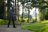 Persona, kas valkā drošības aprīkojumu, pļauj zāli gar celiņu ar akumulatora zāles trimmeri.