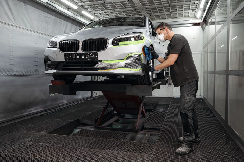Persona, kas valkā drošības aprīkojumu, slīpē automašīnas buferi ar akumulatora ekscentrisko slīpmašīnu.