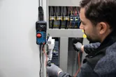 Persona, kas valkā drošības aprīkojumu, izmanto digitālo multimetru, lai pārbaudītu elektrisko paneli.