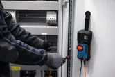 Persona, kas valkā drošības aprīkojumu, izmanto digitālo multimetru, lai pārbaudītu elektrisko paneli.