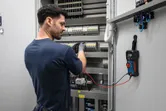 Persona, kas valkā drošības aprīkojumu, izmanto digitālo multimetru, lai pārbaudītu elektrisko paneli.