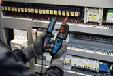 Persona, kas valkā drošības aprīkojumu, izmanto sprieguma testeri uz elektriskā paneļa.