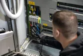 Persona, kas valkā drošības aprīkojumu, izmanto sprieguma testeri elektrības paneļa elektroinstalācijai.